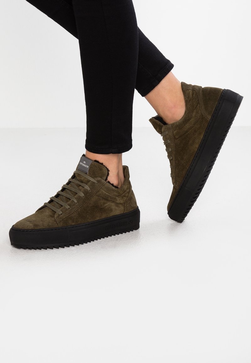 Person trägt olivgrüne Wildledersneaker mit schwarzen Sohlen und schwarze Hose, ein Fuß ruht auf dem anderen vor weißem Hintergrund.