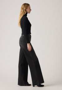 Melna turtlenecks krekls apvienots ar augstā jostasvietā, plašām melnām džinsām ar gludu tekstūru un minimālistisku dizainu. Melni bloka papēžu apavi.