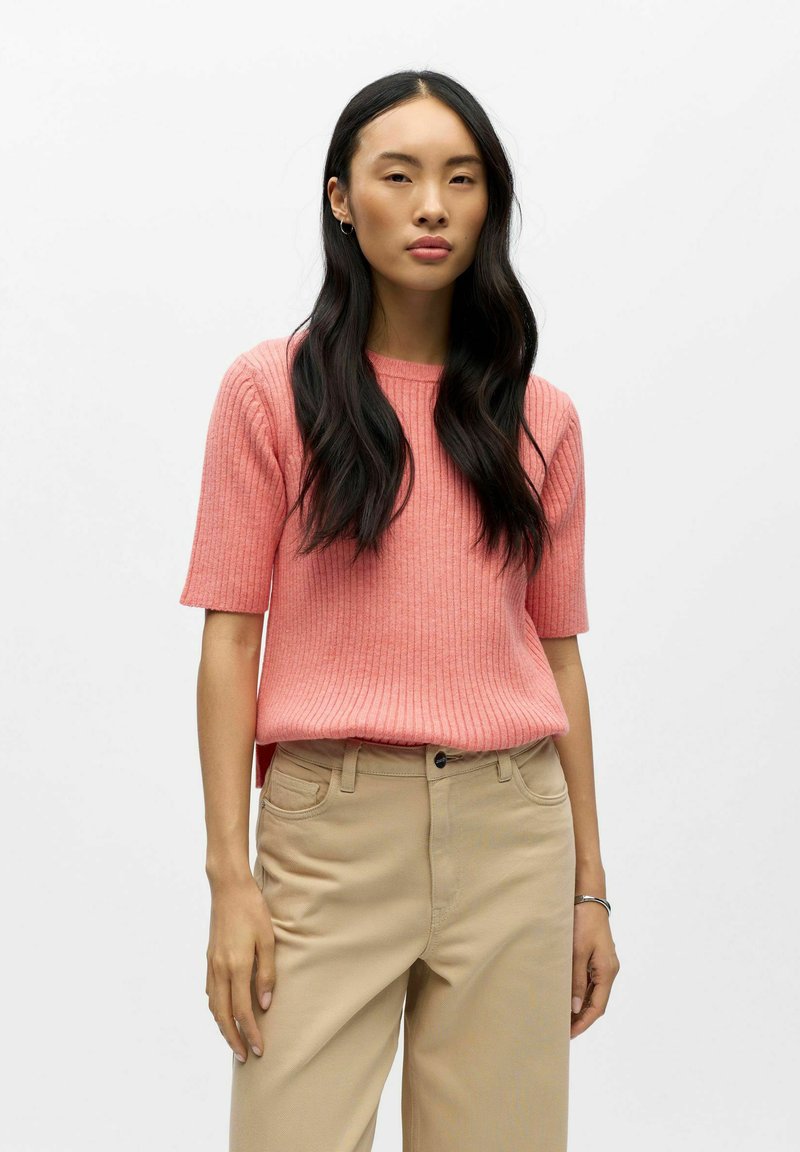 Kortärmelig gebreide top in koraal met een geribbelde textuur en een ronde hals, gecombineerd met licht beige broek met wijde pijpen. Eenvoudig, casual ontwerp.