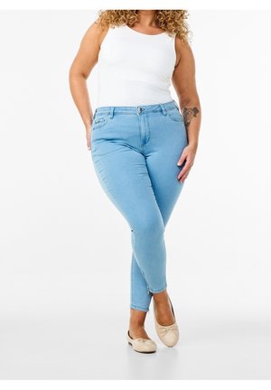 Jeans skinny azzurri chiaro con una texture di denim liscia, dotati di chiusura frontale con bottone e cerniera. Abbinati a una canotta bianca e ballerine beige.