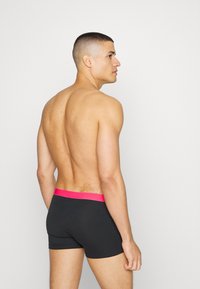 Black boxer shorts feature a bright pink elastic waistband. The model is shown from a side angle, displaying a smooth texture and fitted design.
