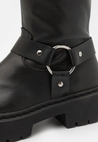 Bota de piel negra hasta el tobillo con una correa ancha y un anillo de metal circular con detalles en plata, suela gruesa con textura de banda.