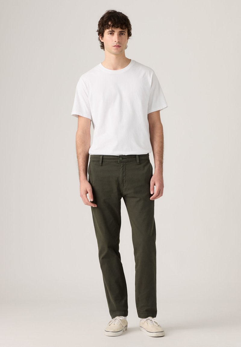 Jeune homme portant un t-shirt blanc uni, un pantalon vert olive et des baskets blanches, debout sur un fond neutre.