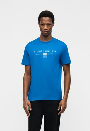 Moški, ki nosi modro majico Tommy Hilfiger z belim logotipom, stoji pred enobarvnim svetlo sivim ozadjem.