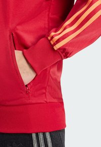 Rote Sportjacke mit einem Reißverschluss-Tasche, die drei horizontale orangefarbene Streifen an den Ärmeln und gerippte Bündchen und Saum aufweist.