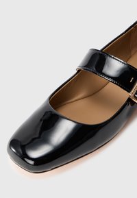 Zapato Mary Jane de charol negro con punta cuadrada y una tira sujeta por una hebilla dorada sobre una plantilla marrón, sobre fondo blanco.