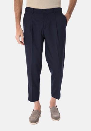 Uomo che indossa pantaloni blu navy plissettati e corti con scarpe beige slip-on, in piedi con una mano in tasca contro uno sfondo semplice.