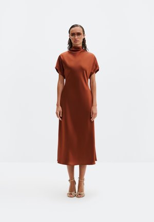 Kobieta z mokrymi włosami, ubrana w pomarańczową sukienkę midi z wysokim kołnierzem, okulary przeciwsłoneczne oraz beżowe sandały na obcasie, stojąca na białym tle.