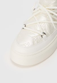 Zapatillas de deporte blancas de piel sintética con parte superior texturizada, costuras tonales y cordones planos blancos. Presenta una gruesa suela de goma blanca.
