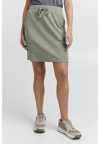 Fustă verde deschis, cu nervuri, dotată cu un brâu elastic ajustabil, purtată împreună cu teniși atletici bej și gri. Design simplu și casual.