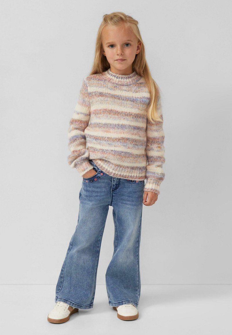 Texturierter Strickpullover in Pastellfarben mit horizontalen Streifen, kombiniert mit hellblauen, weiten Jeans und weißen Sneakers mit braunen Sohlen.