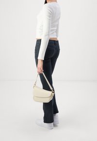 Persona vistiendo una blusa de manga larga blanca y pantalones oscuros de rayas, sujetando un pequeño bolso beige, de pie sobre un suelo blanco con un fondo liso.