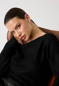 Maglione nero lavorato a maglia con una scollatura ampia e maniche a costine. La modella ha i capelli lisci, indossa orecchini a cerchio e un sottile anello.