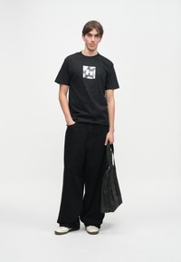 Νέος/νέα με κοντά μαλλιά, φορώντας μαύρο μπλουζάκι με γραφικά και φαρδιά μαύρα παντελόνια, κρατώντας μια μαύρη τσάντα tote, που στέκεται μπροστά σε λευκό φόντο.