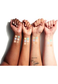 Trois bras de différentes teintes de peau présentant des échantillons de diverses teintes de fond de teint, y compris beige, marron et des couleurs pastel.