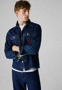Chaqueta de denim en azul oscuro con dos bolsillos en el pecho, botones blancos y bordado rojo en el bolsillo izquierdo. Superpuesta sobre una camiseta blanca.