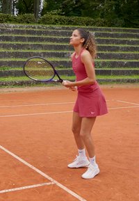 Jucător de tenis îmbrăcat într-o rochie fără mâneca, roz, cu adidași albi, pregătindu-se să servească pe un teren de zgură, cu arbuști verzi în fundal.