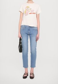 Kremowa koszulka z nadrukiem graficznym cheruba i napisem, zestawiona z jasnoniebieskimi dżinsami, czarną torebką i brązowymi płaskimi butami z paskami.