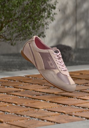 Sneaker rosa chiaro e beige con lacci che fluttuano sopra una superficie piastrellata in legno e pietra all'aperto vicino a un muro e una pianta verde.