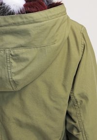 Oliwkowa kurtka z kapturem z miękkim, teksturowanym kołnierzem. Ma luźny krój i widoczne szwy na plecach.