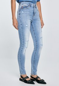 Inimene, kes kannab helesiniseid rebenenud kitsaid teksaseid ja musti terava ninaga rihmlukke sisaldavaid plätusid, seisab ühevärvilise tausta ees.