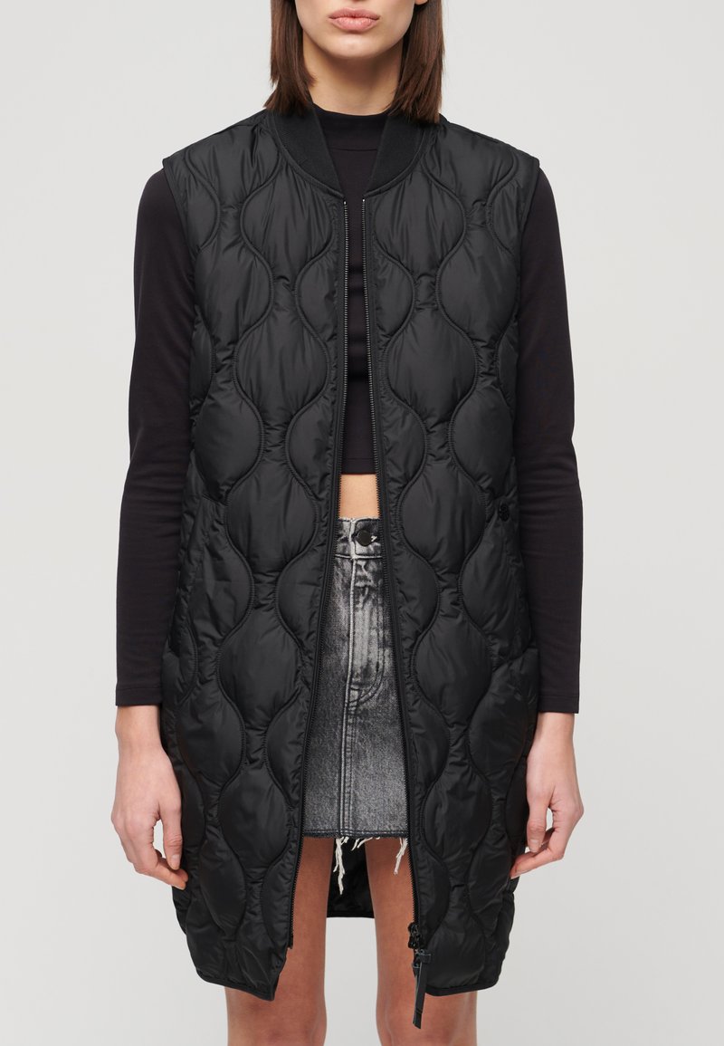 Schwarze gepolsterte ärmellose Weste mit runder Steppung, Frontreißverschluss und geripptem Kragen, kombiniert mit einem schwarzen Oberteil und einem Jeansrock.