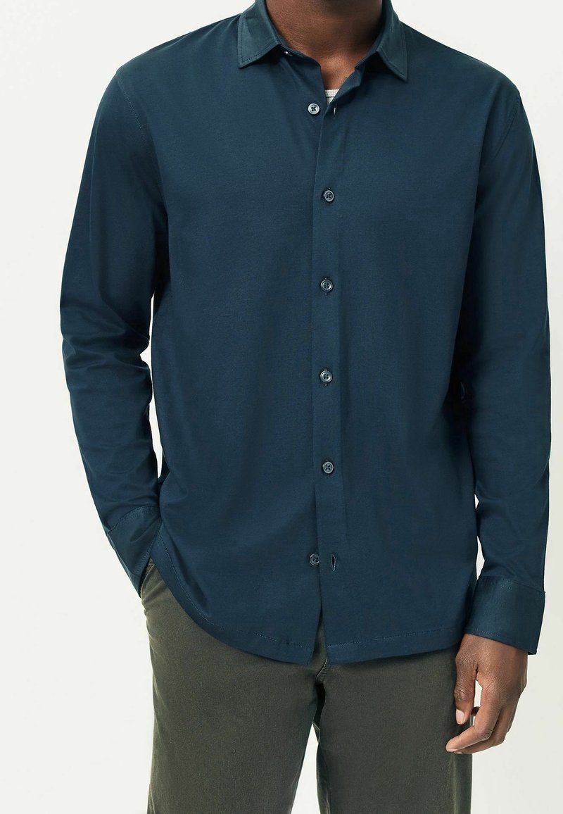 Homme portant une chemise bleu foncé à manches longues boutonnée et un pantalon vert foncé, une main dans la poche, debout devant un fond clair.
