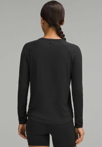 Crna sportska majica dugih rukava s teksturiranim završetkom, raglan rukavima i malim detaljem na zatvaranju na leđima; uparen s crnim šortsem.