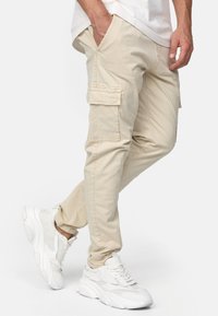 Beige cargo-housut, joissa on sivutaskut, kapea istuvuus ja kevyt kangas. Yhdistetty valkoisiin urheilukengiin.