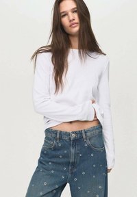 White long-sleeve shirt paired with high-waisted blue jeans featuring small circular embroidered details. Casual fit, soft fabric, relaxed design.