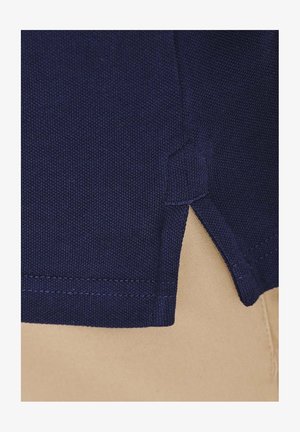 Granatowy, teksturowany materiał z rozcięciem u dołu. Szw nadchodzą drobno, kontrastując z jasnobeżowym tłem.