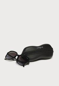 Ochelari de soare negri cu o ramă groasă, lentile gradient și accente aurii, alături de un toc negru cu fermoar care prezintă logo-ul "vogue."
