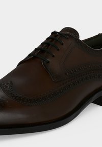 Chaussure habillée en cuir marron foncé pour homme avec détails brogue et lacets noirs sur fond blanc.