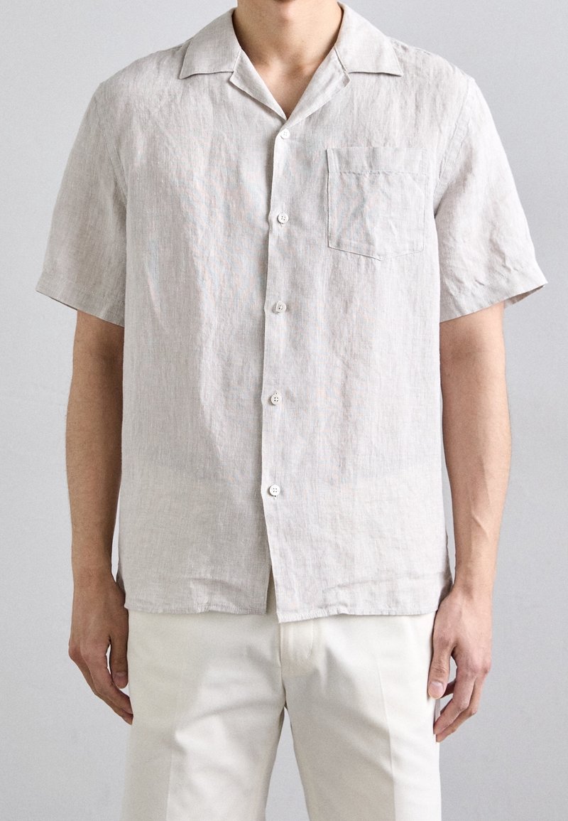 Homme portant une chemise beige clair à manches courtes avec une poche poitrine et un pantalon blanc cassé sur un fond uni.