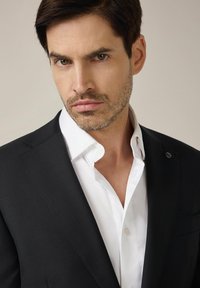 Czarna marynarka na białej koszuli z guzikami, z wysmukłym kołnierzykiem i widocznym guzikiem. Gładki materiał o dopasowanym kroju.