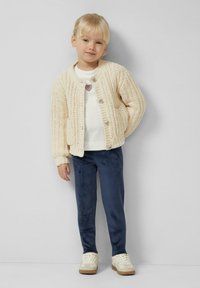 Beige knitted cardigan with ribbed edges, white sweater with heart detail, navy trousers, and off-white sneakers with textured accents.