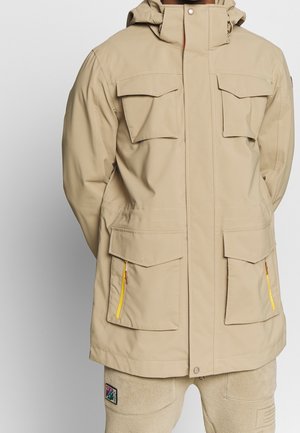 Beżowa kurtka z kapturem z przednimi kieszeniami na patki i żółtymi detalami na suwaku, noszona z beżowymi spodniami z kolorową naszywką na udzie.