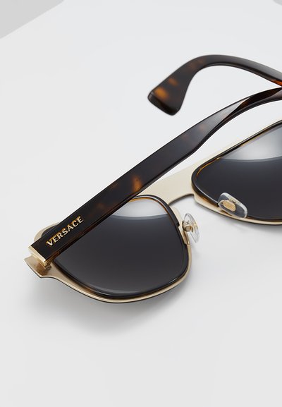 Versace-solbriller med en dristig gullramme, mørke linser og skilpaddebrune tempelarmer. Har klare nesepader og fremtredende merkevare.