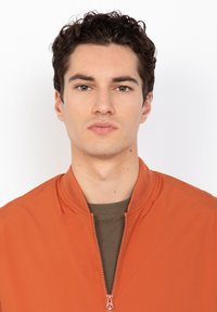 Junger Mann mit dunklem, lockigem Haar, der eine orange Jacke mit Reißverschluss über einem olivgrünen Hemd trägt, vor einem schlichten weißen Hintergrund.