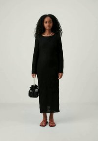 Ilga melna adīta kleita ar garām piedurknēm, apaļu kakla izgriezumu un sarežģītu tekstūru. Apvienota ar melnu somu ar ziedu akcentiem un sarkaniem sandalēm.