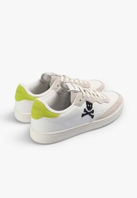 Un par de zapatillas blancas con acentos beige, pestañas en el talón de color verde lima, diseño de calavera y tibias cruzadas en los lados, y cordones blancos, en ángulo a la derecha.