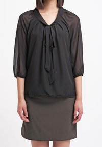 Zwarte, sheer blouse met driekwartmouwen en een strikdetail bij de hals, gecombineerd met een aangesloten olijfgroene rok. Gladde textuur, minimalistisch design.