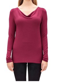 Kobieta w długim, dopasowanym, ciemnoróżowym swetrze z szerokim, luźno opadającym dekoltem oraz czarnych spodniach, stoi z rękami swobodnie wzdłuż ciała.