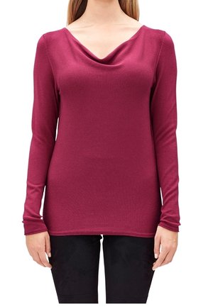 Frau trägt einen langärmeligen, taillierten, tiefrosa Pullover mit einem weiten, drapierter Ausschnitt und schwarzen Hosen, steht mit entspannten Armen an den Seiten.