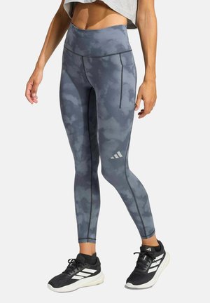 Frau trägt hochtaillierte graue Camouflage-Leggings mit Seitentasche und schwarze Laufschuhe mit weißen Streifen, steht vor weißem Hintergrund.