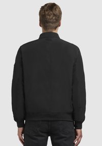 Schwarze Bomberjacke mit geripptem Kragen und Manschetten, die eine glatte Textur und keine sichtbaren Muster aufweist. Rückansicht mit minimaler Detailgestaltung.