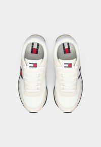 Sneakers bianchi e beige con design a lacci, caratterizzati da strisce rosse e blu navy, e un patch logo sulla linguetta. Suola in gomma testurizzata.