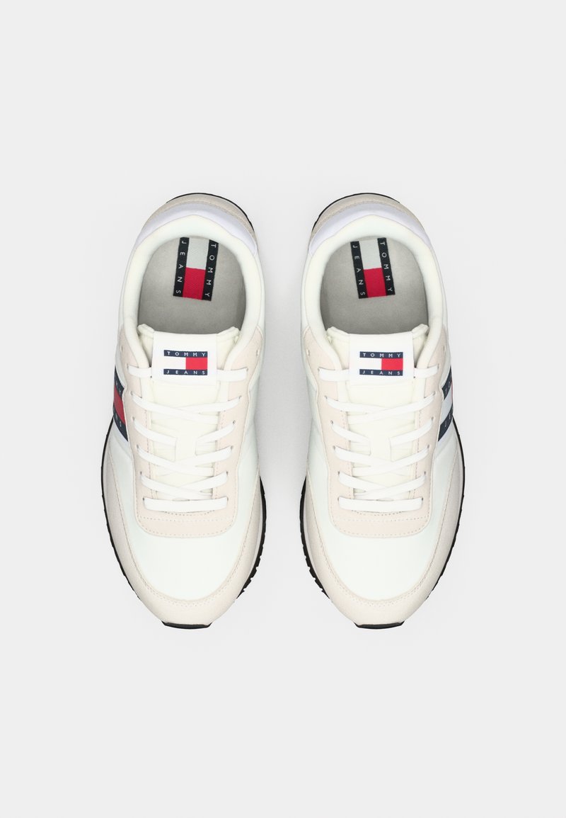 Zapatillas blancas y beige con diseño de cordones, que presentan rayas rojas y marinas, y un parche con el logo en la lengüeta. Suela de goma texturizada.