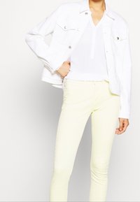 Giacca in denim bianco abbinata a una camicia bianca a scollo a V e pantaloni slim-fit giallo pallido. La giacca presenta due tasche sul petto e dettagli con bottoni metallici.
