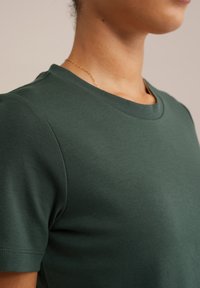 Zeleni bombažni majica s kratkimi rokavi in okroglim izrezom, gladke teksture in enostavnega dizajna. Ima dvojno šivano rob za trajnost.